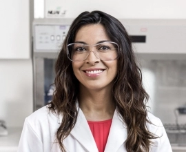 A female Novartis scientist wearing a white lab coat and glasses, smiles in front of laboratory equipment. A female Novartis scientist wearing a white lab coat and glasses, smiles in front of laboratory equipment.